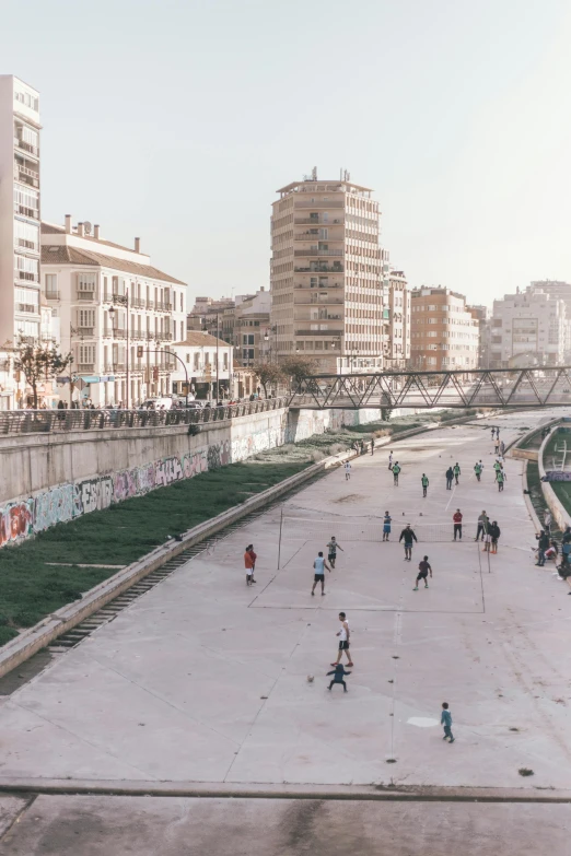 a group of people walking down a street next to a river, inspired by Oswaldo Viteri, street art, playing soccer, panoramic view, 2022 photograph, skatepark