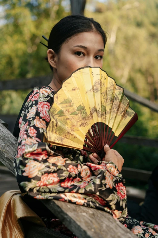 a woman sitting on a bench holding a fan, inspired by Fan Kuan, unsplash, renaissance, wearing yellow floral blouse, black, mulan, brown