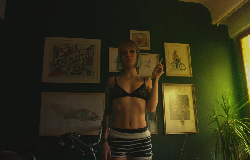 a woman standing in front of a window holding a cigarette, inspired by Elsa Bleda, feminist art, sport bra and dark blue shorts, in a cyberpunk themed room, movie frame still, with tattoos