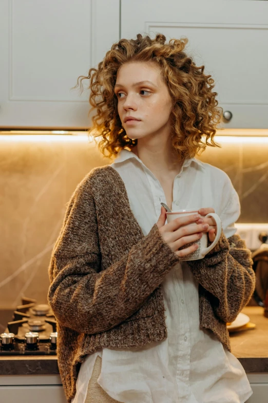 a woman standing in a kitchen holding a cup, by Alexander Brook, trending on pexels, renaissance, brown sweater, curly haired, muted lights, teenager girl