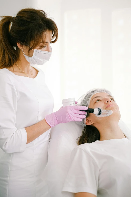 a woman getting a facial mask at a beauty salon, surgical gear, woman holding another woman, with a white background, blushing