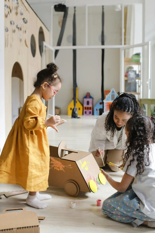 two little girls are playing with a cardboard box, pexels contest winner, interactive art, lady using yellow dress, in a workshop, inspect in inventory image, made of cardboard