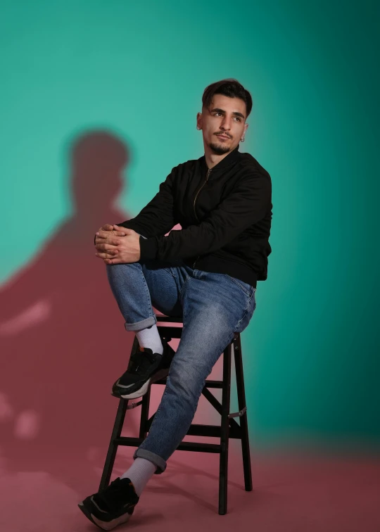 a man sitting on top of a wooden chair, by William Berra, trending on reddit, teal studio backdrop, bisexual lighting, wearing jeans and a black hoodie, art foreground : eloy morales