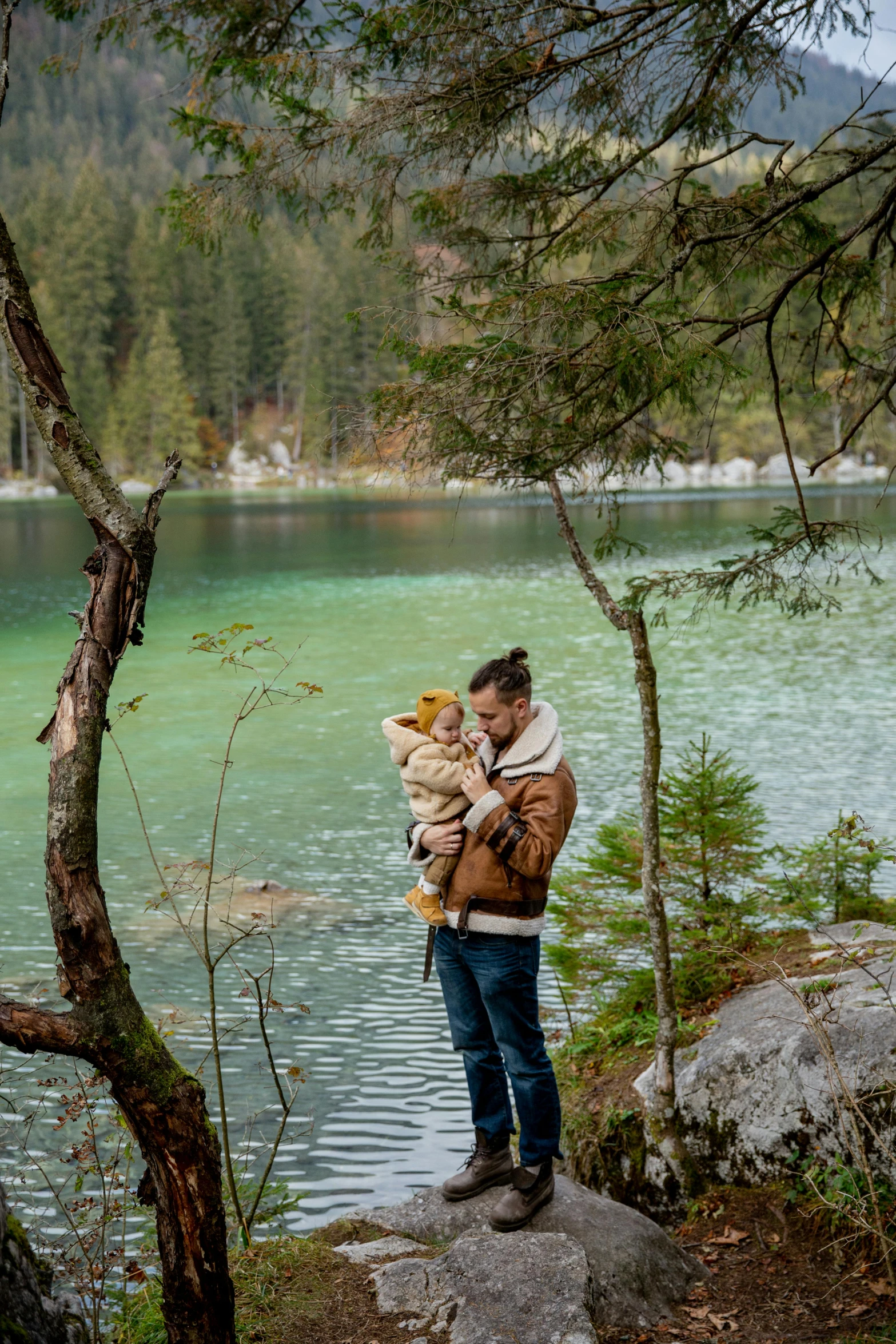 a man standing on top of a rock next to a lake, by Julia Pishtar, holding a boxer puppy, in forest, with a kid, cosy atmoshpere