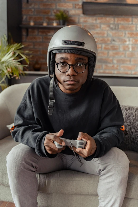 a man sitting on a couch holding a video game controller, inspired by Xanthus Russell Smith, trending on pexels, afrofuturism, wearing a helmet, wearing black frame glasses, wearing a track suit, gray men