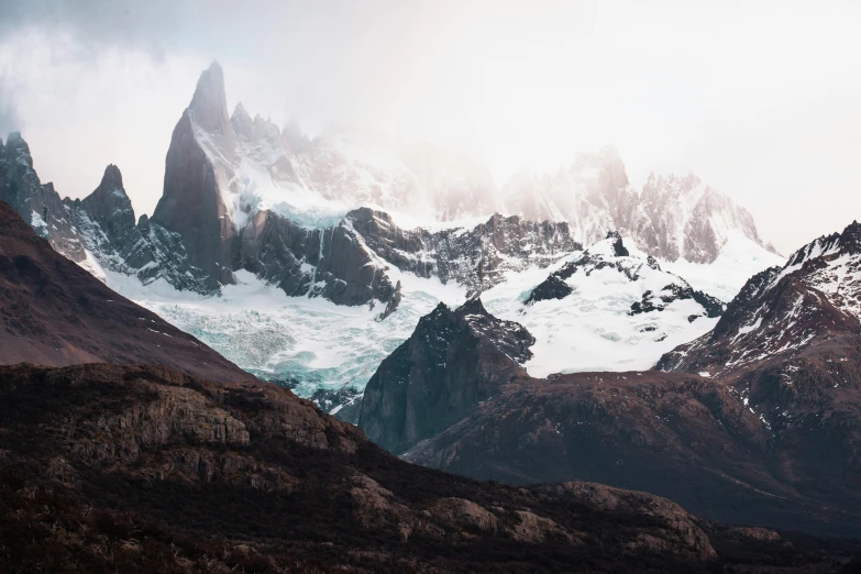 a group of mountains covered in snow on a cloudy day, a matte painting, by Lucas Vorsterman, unsplash contest winner, patagonian, megan fox as beautiful mountains, tall stone spires, extreme panoramic