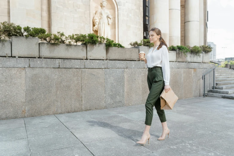 a woman walking down a sidewalk next to a building, olive green slacks, celebration of coffee products, wearing an elegant outfit, milk and mocha style