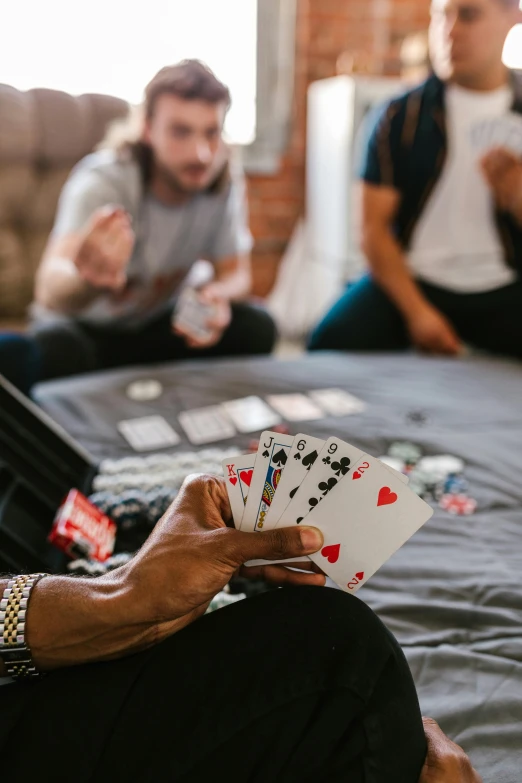 a man sitting on top of a bed holding playing cards, a screenshot, by Adam Marczyński, pexels contest winner, families playing, dave chappelle, laying down with wrists together, playboi carti