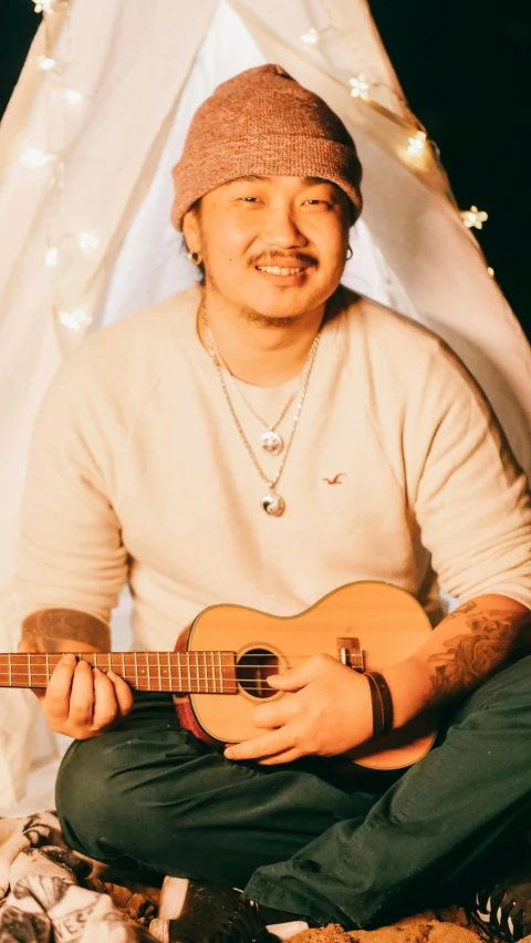 a man sitting in front of a teepee with a guitar, an album cover, inspired by Eddie Mendoza, unsplash, dau-al-set, asian male, coy smile, mr beast, studio picture