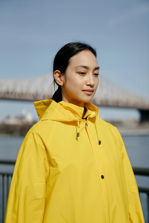 a woman standing in front of a bridge, yellow raincoat, sleek waterproof design, greta thunberg, kakar cheung