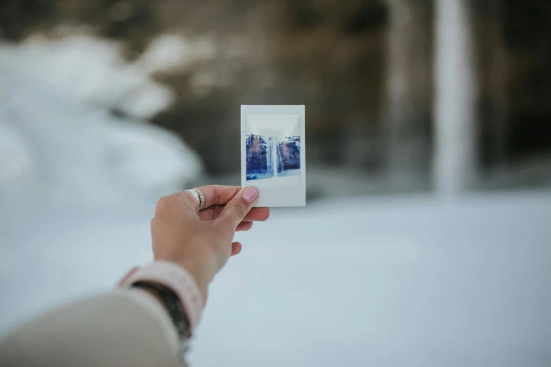 a person holding up a polaroid picture in the snow, by Emma Andijewska, pexels contest winner, visual art, looking outside, holding flask in hand, vertical orientation, symmetrical shot