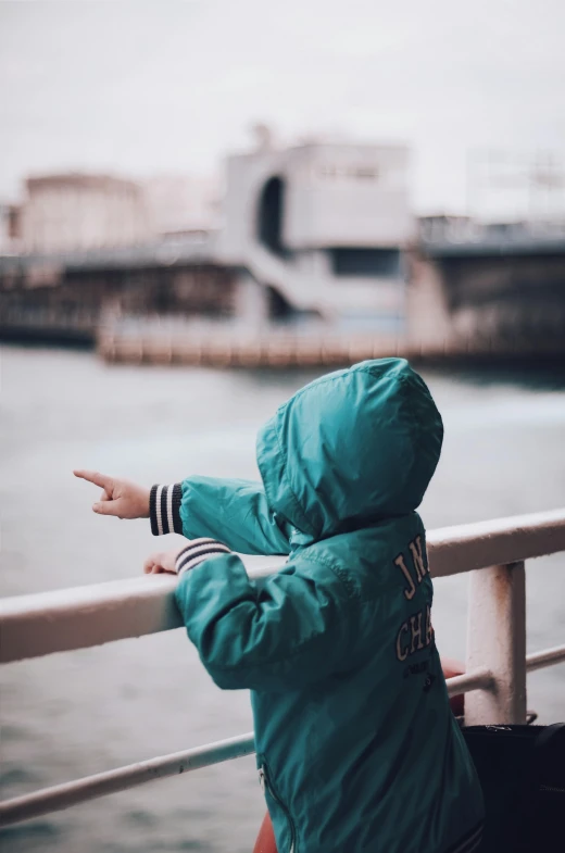 a person standing next to a body of water, little kid, with teal clothes, looking into the distance, pointing