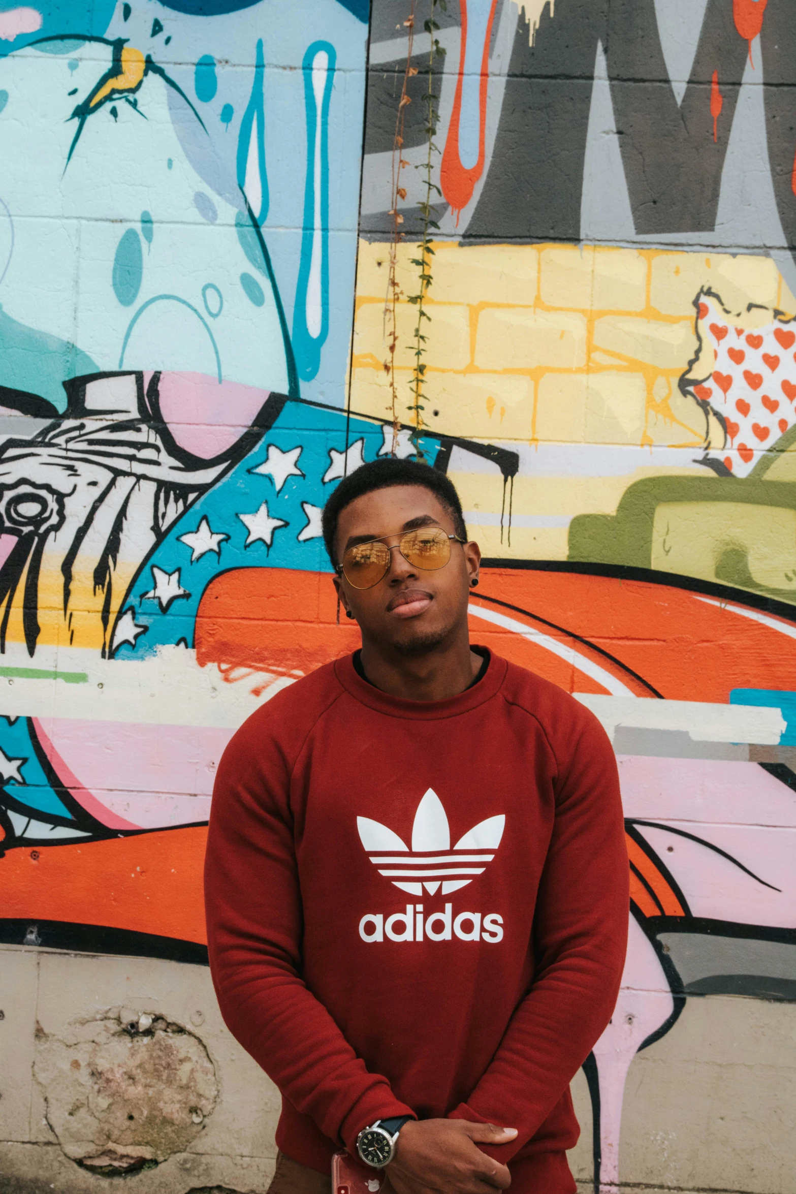 a man standing in front of a wall covered in graffiti, an album cover, by Austin English, trending on unsplash, wearing adidas clothing, he is wearing a brown sweater, in style of tyler mitchell, promotional image