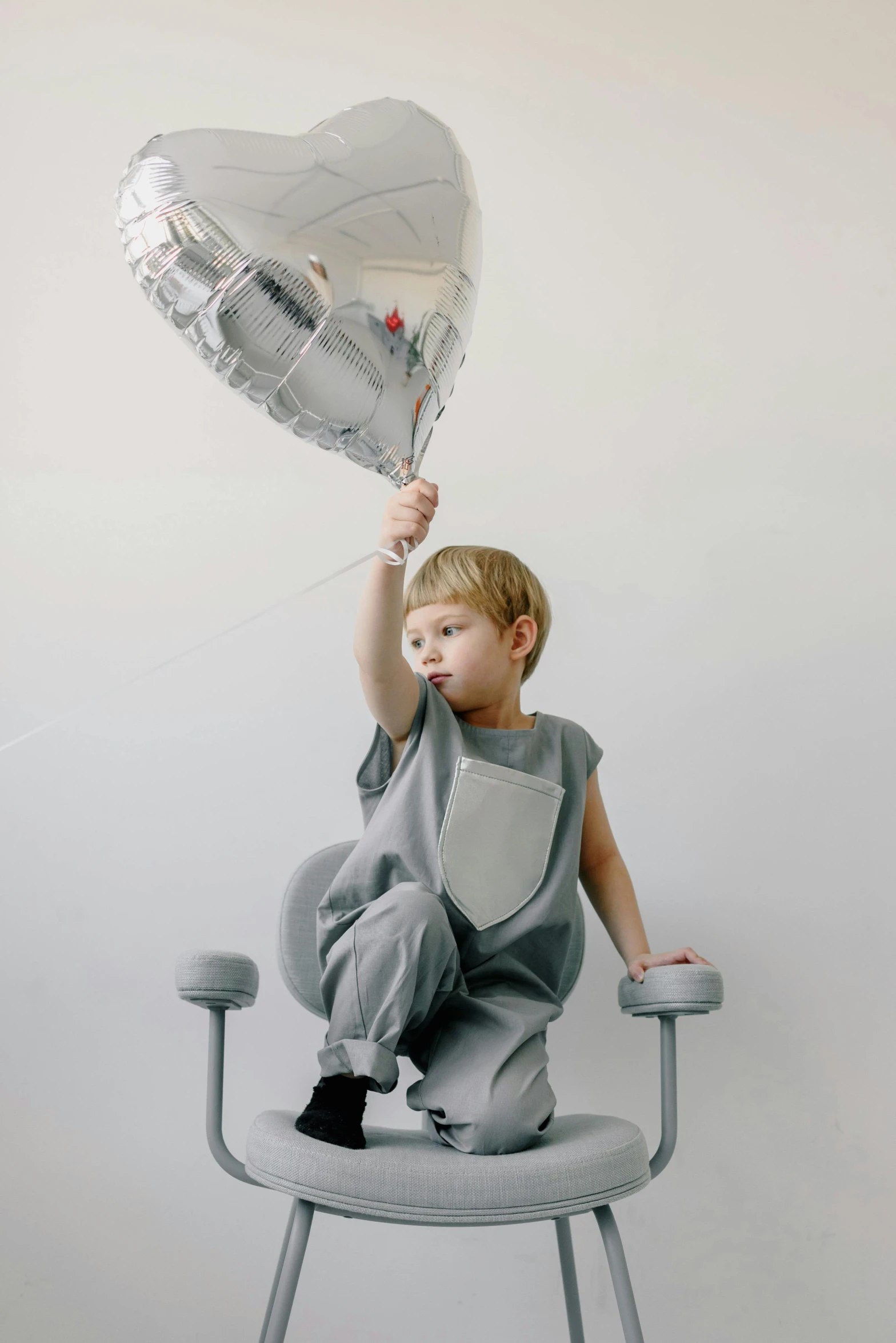 a little boy sitting in a chair holding a heart shaped balloon, unsplash, silver garment, wearing human air force jumpsuit, light grey, sitting on top of a cryopod