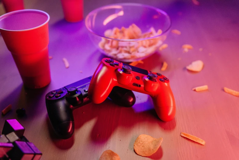 a video game controller sitting next to a bowl of chips, a still life, by Julia Pishtar, pexels, orange and red lighting, purple and red, college party, pair of keycards on table