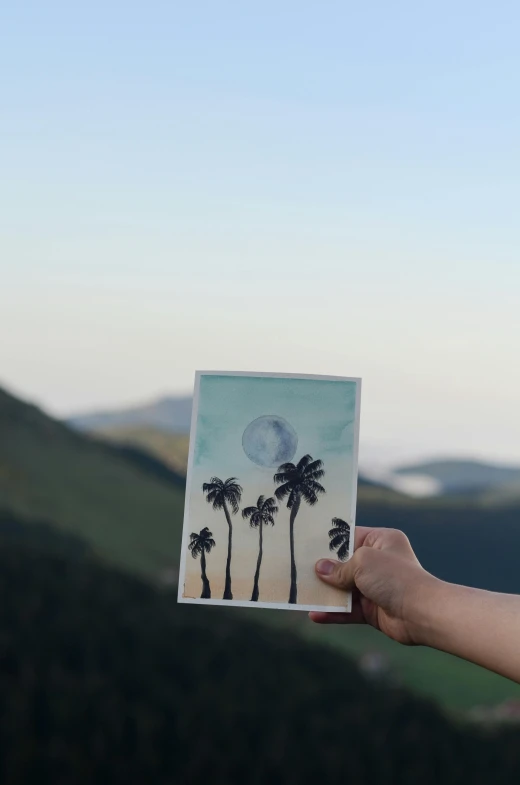 a person holding up a picture of palm trees, pexels contest winner, serial art, in the mountains, big moon on the right, on high-quality paper, high angle shot