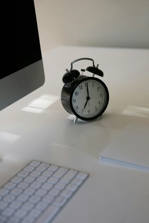 an alarm clock sitting on top of a desk next to a computer, on a white table, looking towards camera, evenly lit, glossy surface