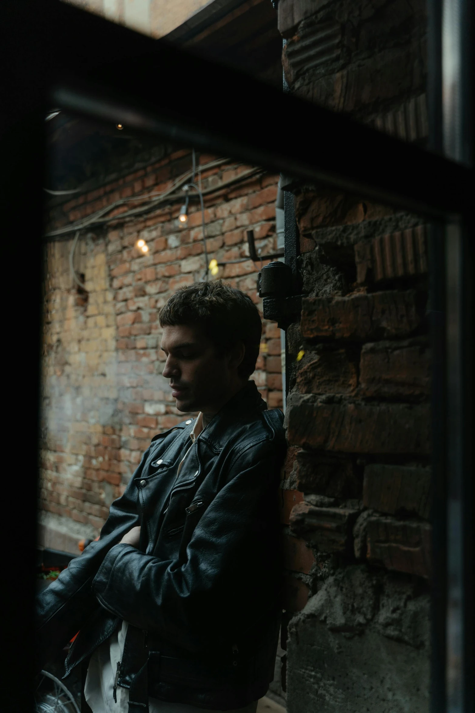 a man standing in front of a window next to a brick wall, inspired by Elsa Bleda, wearing a leather jacket, sophia lillis, cinematic shot ar 9:16 -n 6 -g, sitting alone at a bar