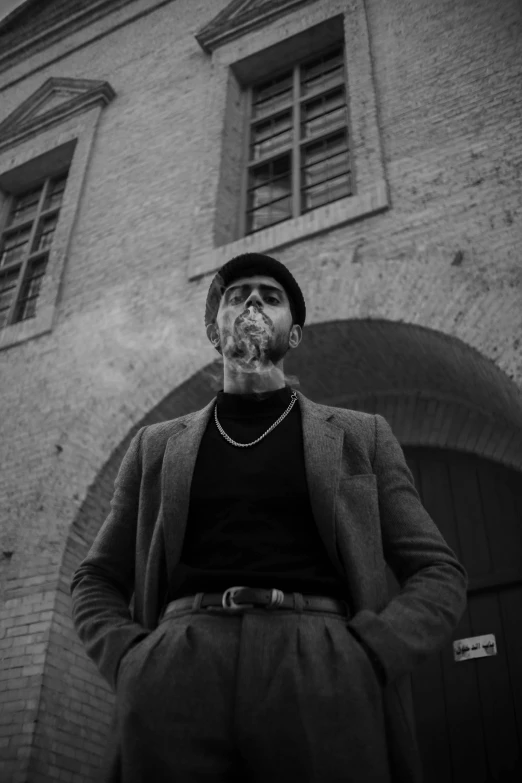 a man with a beard standing in front of a building, a black and white photo, inspired by Theo Constanté, pexels contest winner, cigarette in his mouth, rapper, noir animation, black man