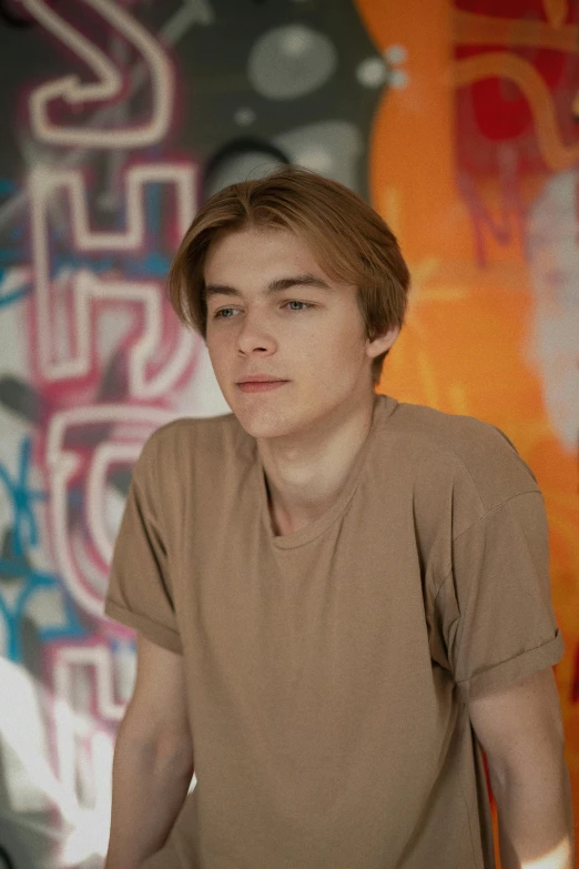 a young man standing in front of a graffiti covered wall, a picture, inspired by Nikolaj Abraham Abildgaard, joe keery, canvas, indoor picture, ansel ]