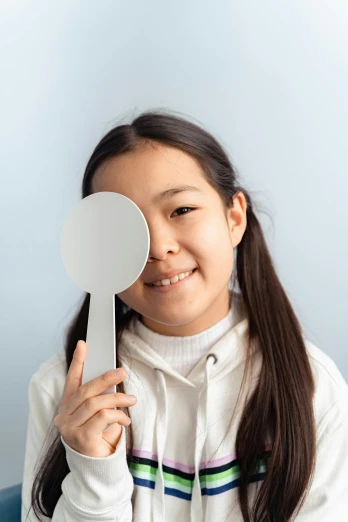 a young girl holding a mirror in front of her face, an album cover, inspired by Kanō Naizen, pexels contest winner, working in her science lab, south east asian with round face, on grey background, optical lenses