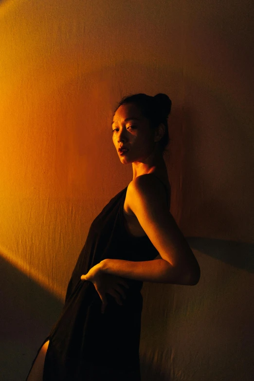 a woman in a black dress leaning against a wall, an album cover, inspired by Gao Xiang, pexels contest winner, sunbathed skin, dramatic lowkey studio lighting, bella poarch, radiating golden light