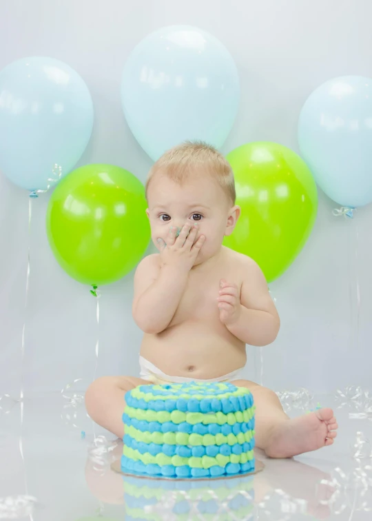 a baby sitting in front of a cake covered in frosting, shutterstock contest winner, green and blue color scheme, baloons, holding his hands up to his face, press shot