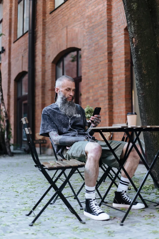 a man sitting at a table next to a tree, pexels contest winner, realism, brown buzzcut, cellphone, greybeard, street clothing