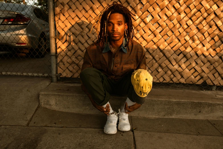 a man with dreadlocks sitting in front of a fence, by Stokely Webster, holding a gold bag, man holding a balloon, sneaker photo, (golden hour)