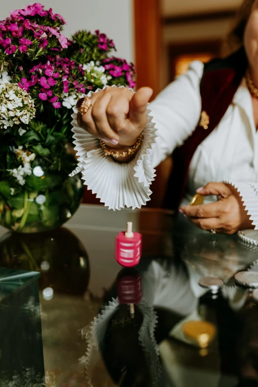 a woman sitting at a table in front of a vase of flowers, coated pleats, carrying a bottle of perfume, anjali mudra, zoomed in shots