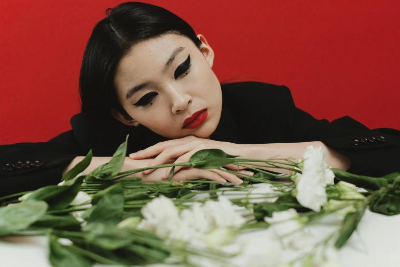 a woman laying on top of a table next to a bunch of flowers, an album cover, inspired by Min Zhen, tumblr, wearing a black and red suit, close up portrait photo, gongbi, on a pale background