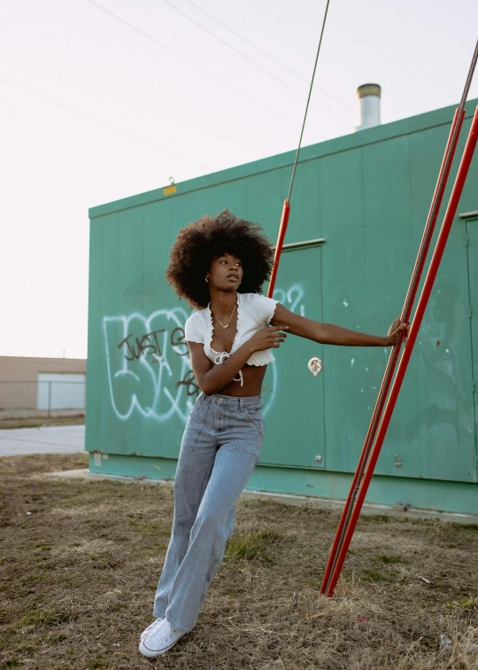 a woman holding a pole in front of a building, an album cover, trending on pexels, black arts movement, black man with afro hair, wearing blue jean overalls, at a fashion shoot, in the yard