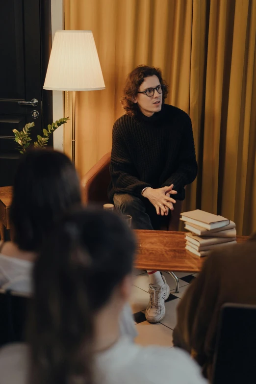 a group of people sitting around a table in a room, he is wearing a brown sweater, julia hetta, on a coffee table, wearing black frame glasses