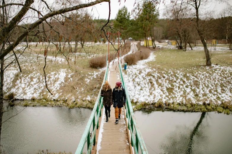 two people walking across a bridge over a river, by Julia Pishtar, pexels contest winner, hurufiyya, outside winter landscape, cardboard, lesbians, well built
