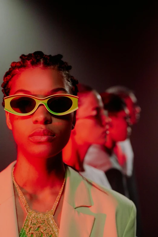a woman standing in front of a group of people wearing sunglasses, trending on pexels, afrofuturism, yellow carrera glasses, studio lit, shades green and red, low dark light