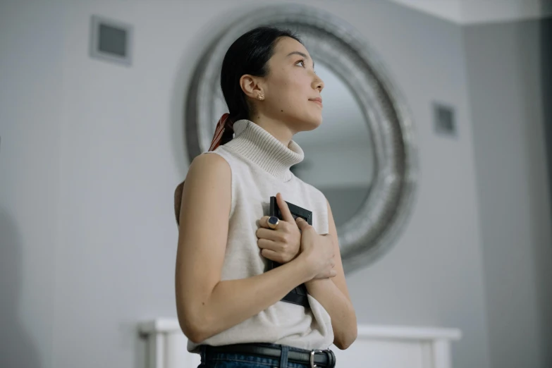 a woman standing in front of a mirror holding a cell phone, inspired by Qian Xuan, pexels contest winner, sleeveless turtleneck, as she looks up at the ceiling, clean and simple, holding a pocket watch