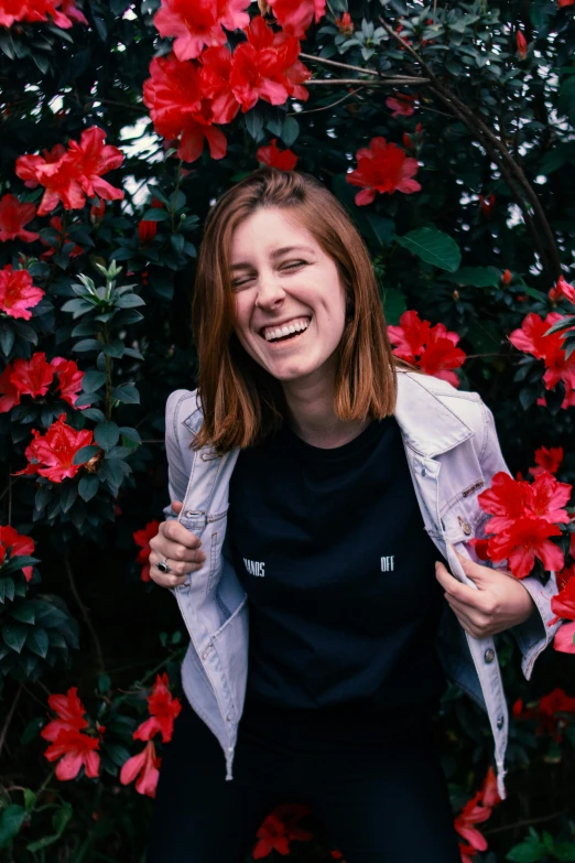 a woman standing in front of a bush of red flowers, a black and white photo, smiling young woman, woman in streetwear, wearing a black jacket, wearing a black tshirt