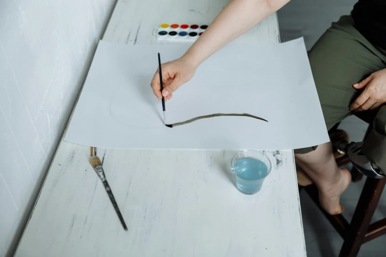a person sitting at a table painting on a piece of paper, a minimalist painting, pexels contest winner, water paint, shaped picture, holding pencil, glass paint