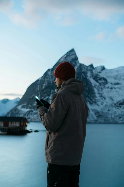 a man standing next to a body of water, by Sebastian Spreng, trending on pexels, holds a smart phone in one hand, icy mountains, looking at spaceships at dock, norwegian man