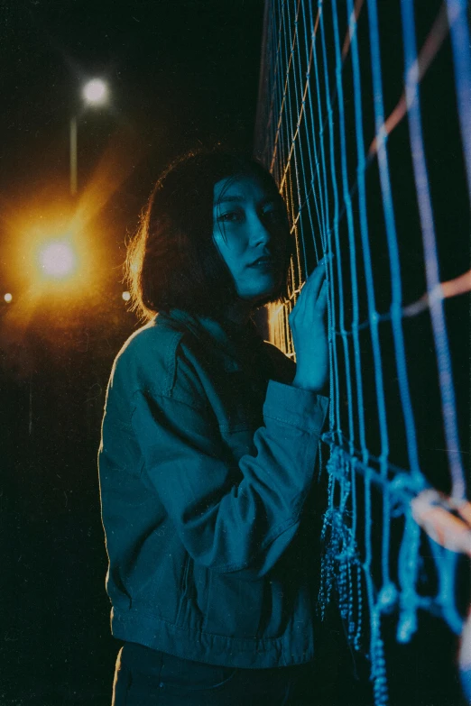 a woman leaning against a fence at night, an album cover, inspired by Elsa Bleda, unsplash, portrait of sadako of the ring, netting, very very low quality picture, looking this way