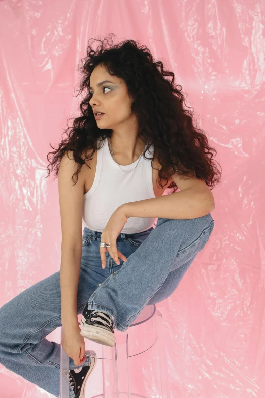 a woman sitting on top of a clear stool, an album cover, trending on pexels, outfit : jeans and white vest, curly haired, hindu aesthetic, she is wearing streetwear