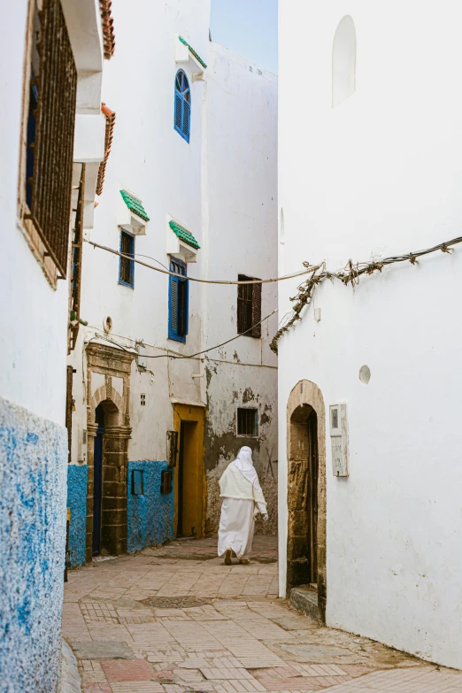 a person in a white robe walking down a narrow street, arabesque, blue and white colour scheme, exterior, desert robe, streets