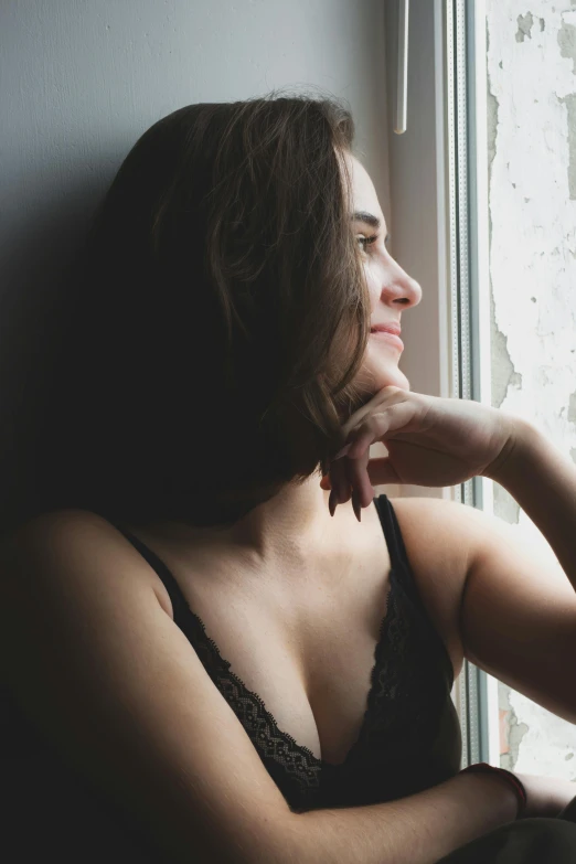 a woman sitting in front of a window looking out, she is wearing a black tank top, profile image, trending photo, shy looking down