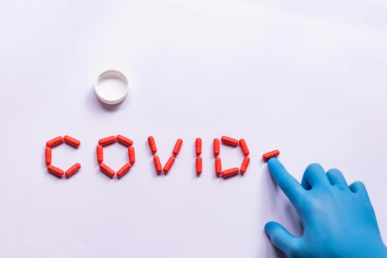 a hand in a blue glove points to the word covidi, by Olivia Peguero, shutterstock, pills, on a white table, covid, red colored