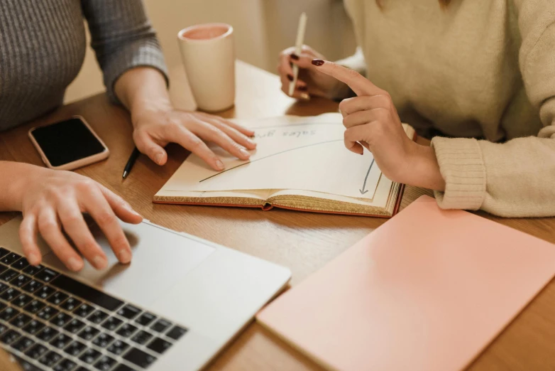 a couple of people sitting at a table with laptops, by Carey Morris, trending on pexels, holding pencil, diary on her hand, background image, character sheets on table