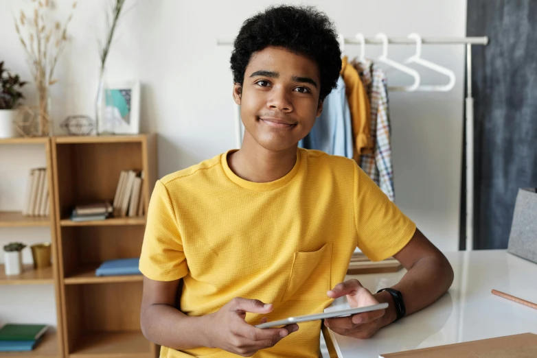 a boy in a yellow shirt holding a cell phone, with brown skin, sitting on a store shelf, in front of a computer, wearing a cute top