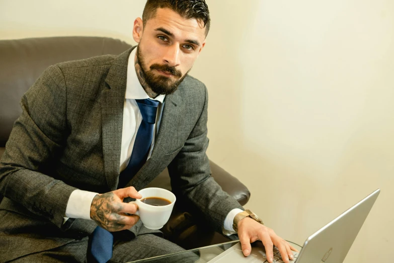 a man sitting in a chair with a laptop and a cup of coffee, wearing a suit and a tie, tattooed man, ash thorp khyzyl saleem, chris cunninham