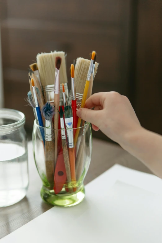 a person holding paint brushes in a glass on a table, our art corner, easy to use, no - text no - logo, full product shot