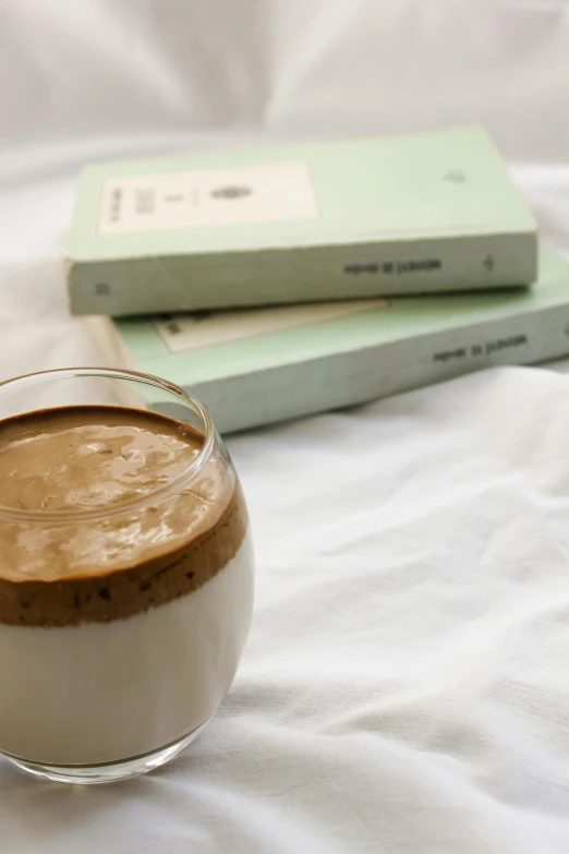 a glass of coffee sitting on top of a bed next to a book, inspired by Richmond Barthé, purism, vanilla smoothie explosion, ignant, chocolate, low - angle shot