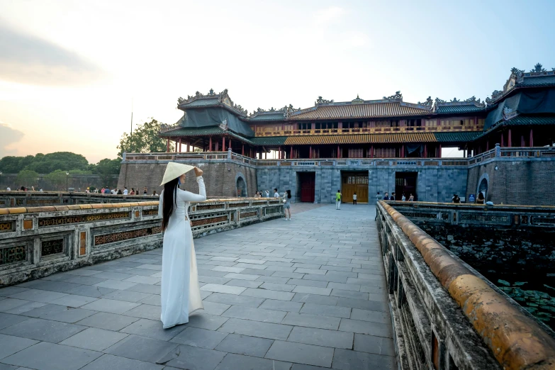 a woman in a white dress standing in front of a building, ao dai, monumental giant palace, wearing a travel hat, square